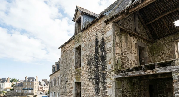 Photographie réaliste d'une maison ou d'un bâtiment ancien montrant des signes d'humidité, de moisissures ou de dommages structurels aux bois (charpente visible, façade abîmée), illustrant le contexte des interventions de rénovation et traitement du bâti menées par 35 ATM dans son rayon d'action. L'image doit montrer un bâtiment typique de la région bretonne en état de dégradation, représentatif des problématiques traitées (humidité, insectes xylophages, état de la couverture).
